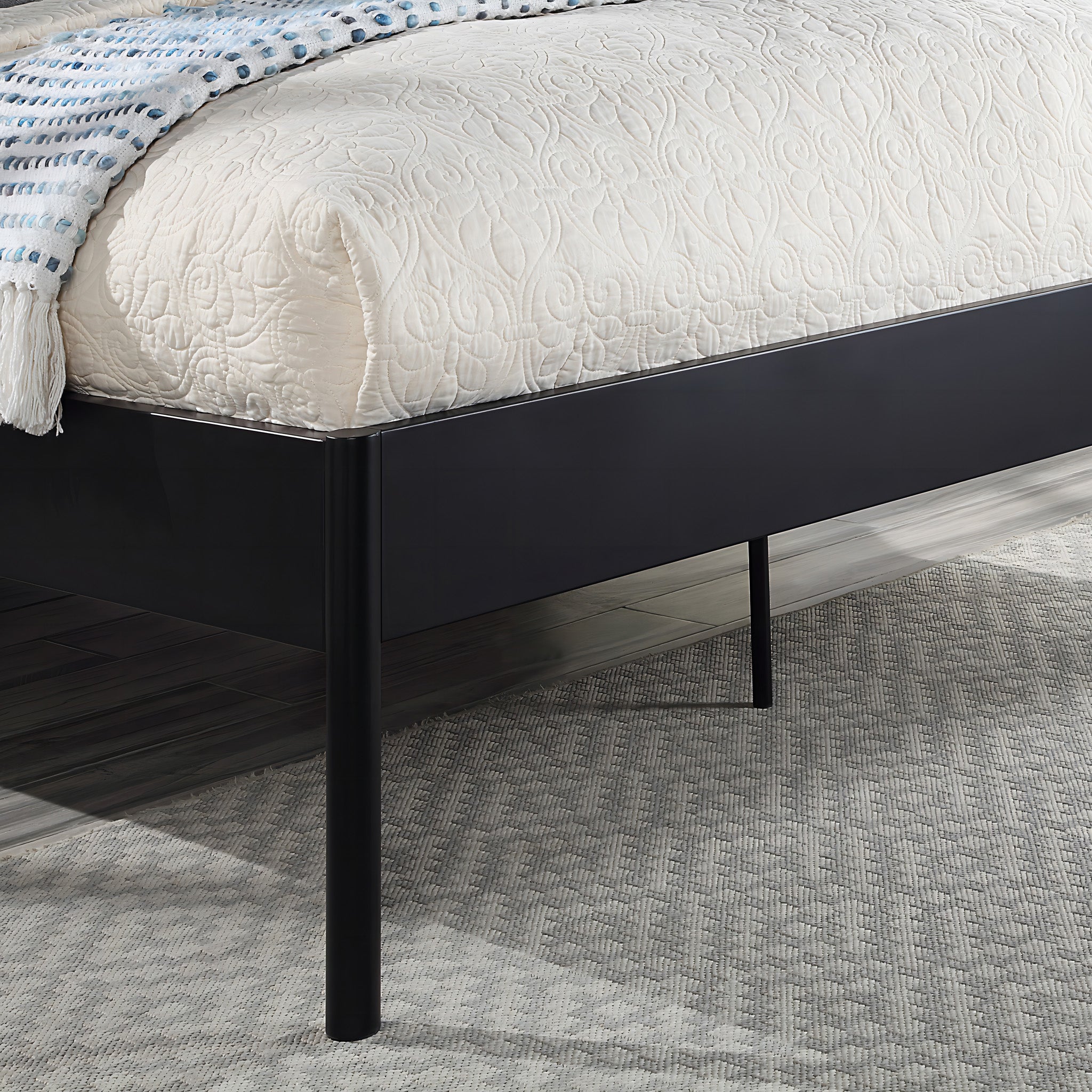 Close-up of the corner of a black metal bed frame with quilted beige bedding and a blue and white throw blanket. The bed is on a light gray carpet, and the floor is dark wood.