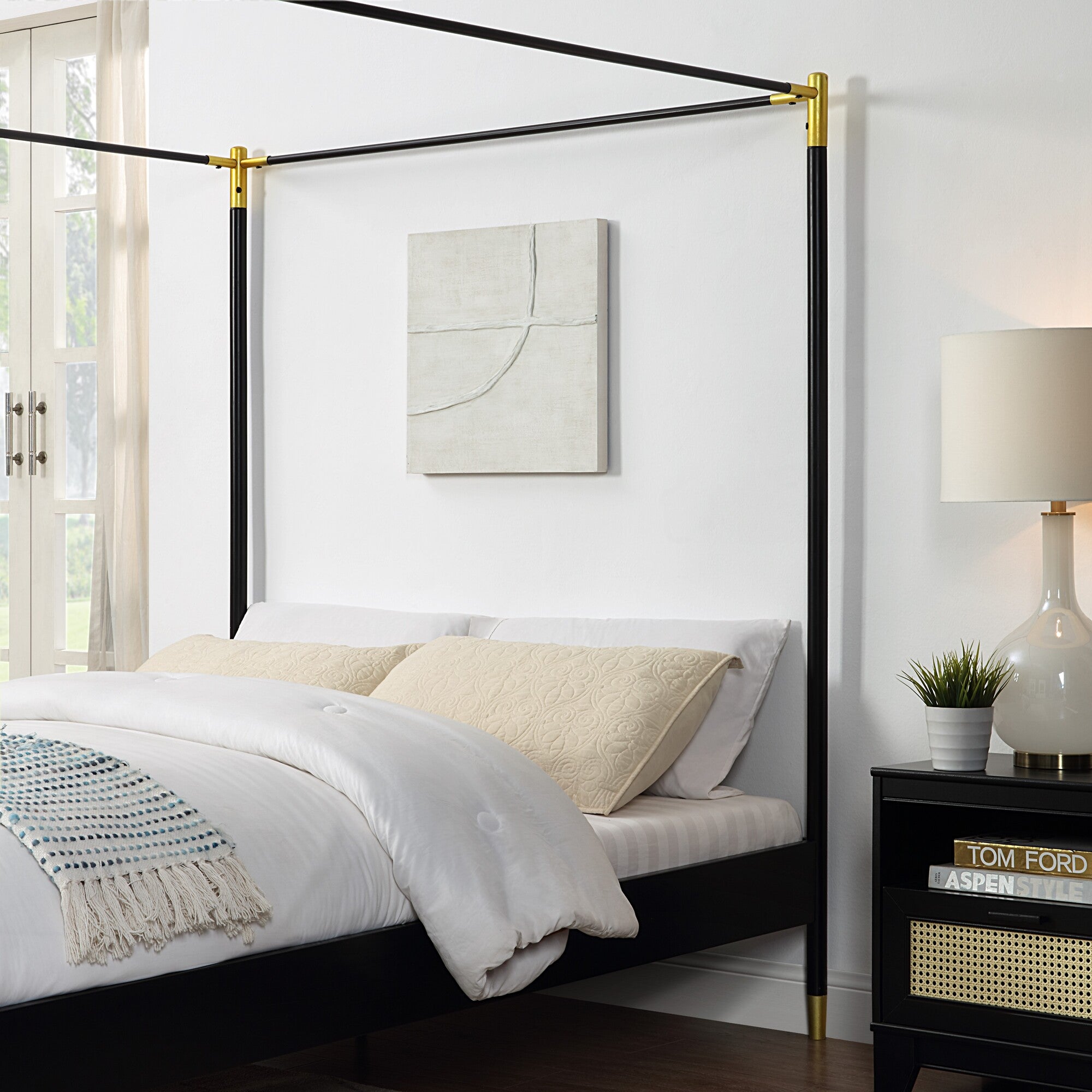 A close-up of a black metal four-poster bed frame with gold accents, set against a white wall, next to a bedside table with a lamp and plant.