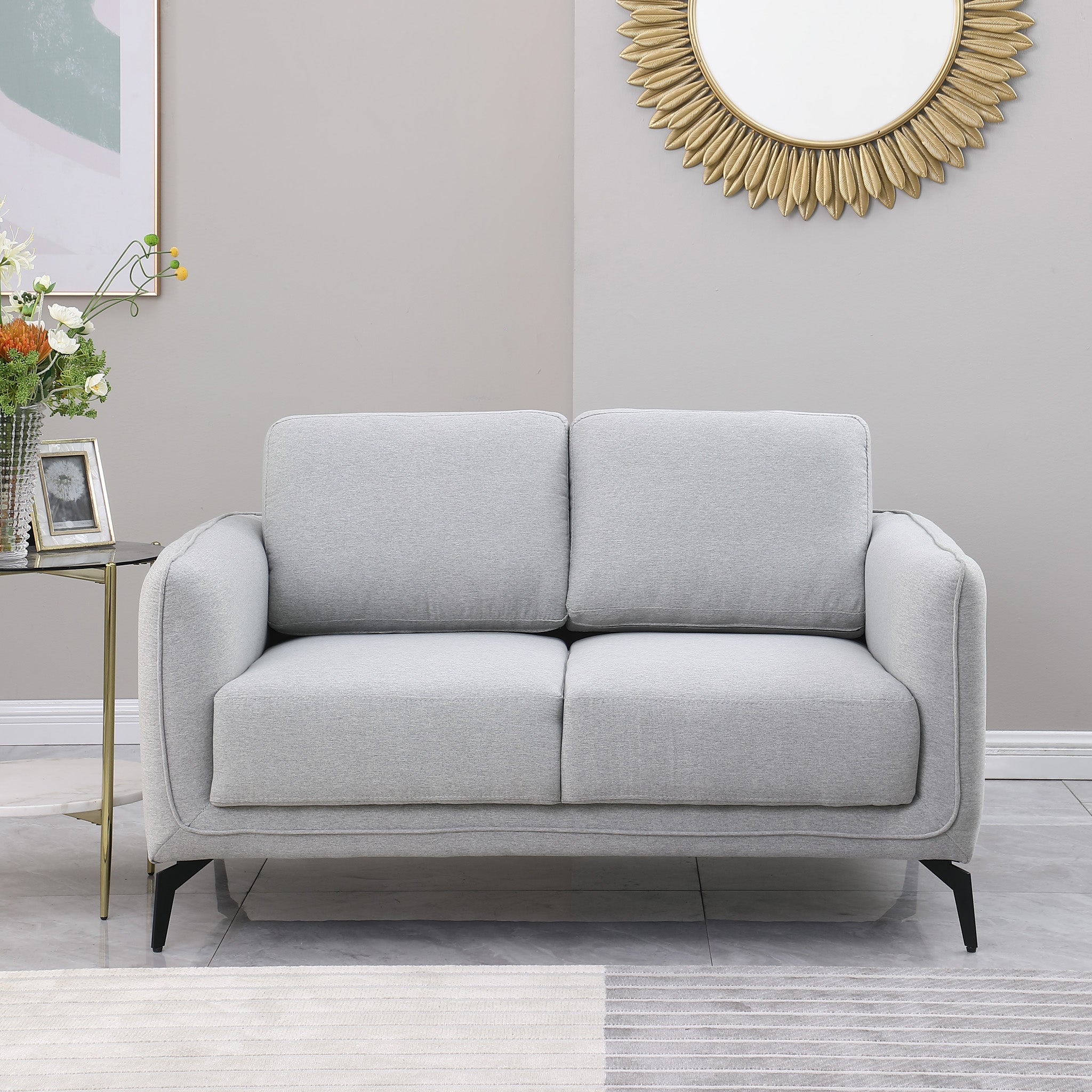 Light grey upholstered sofa with black metal legs in a grey living room, featuring a round sunburst mirror and a side table with flowers.