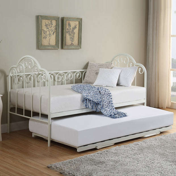 A white metal day bed with an extended trundle is adorned with white bedding, decorative pillows, and a blue throw blanket. Two botanical prints hang above the bed.