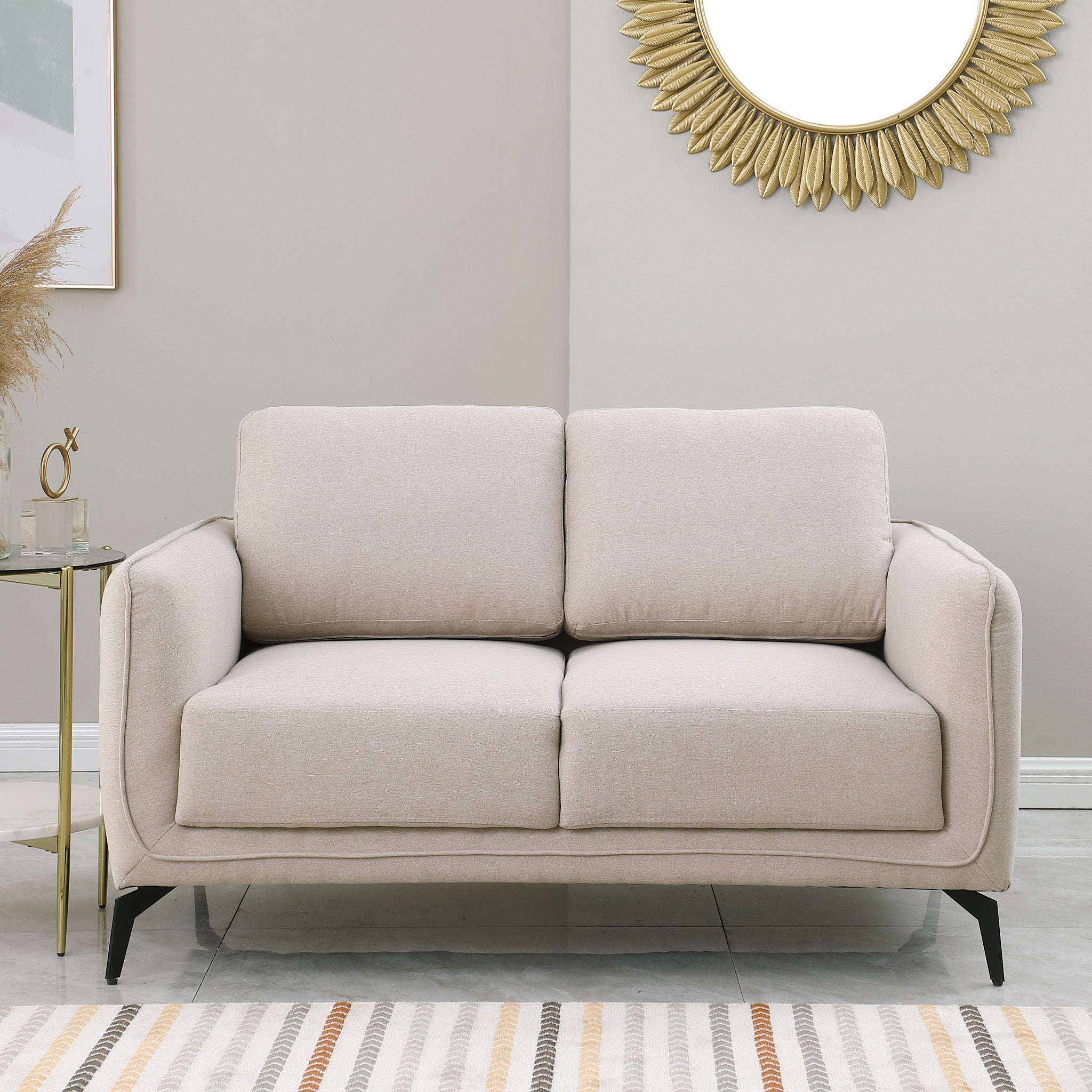 Beige upholstered sofa with black metal legs in a living room, featuring a round sunburst mirror and a side table with flowers.