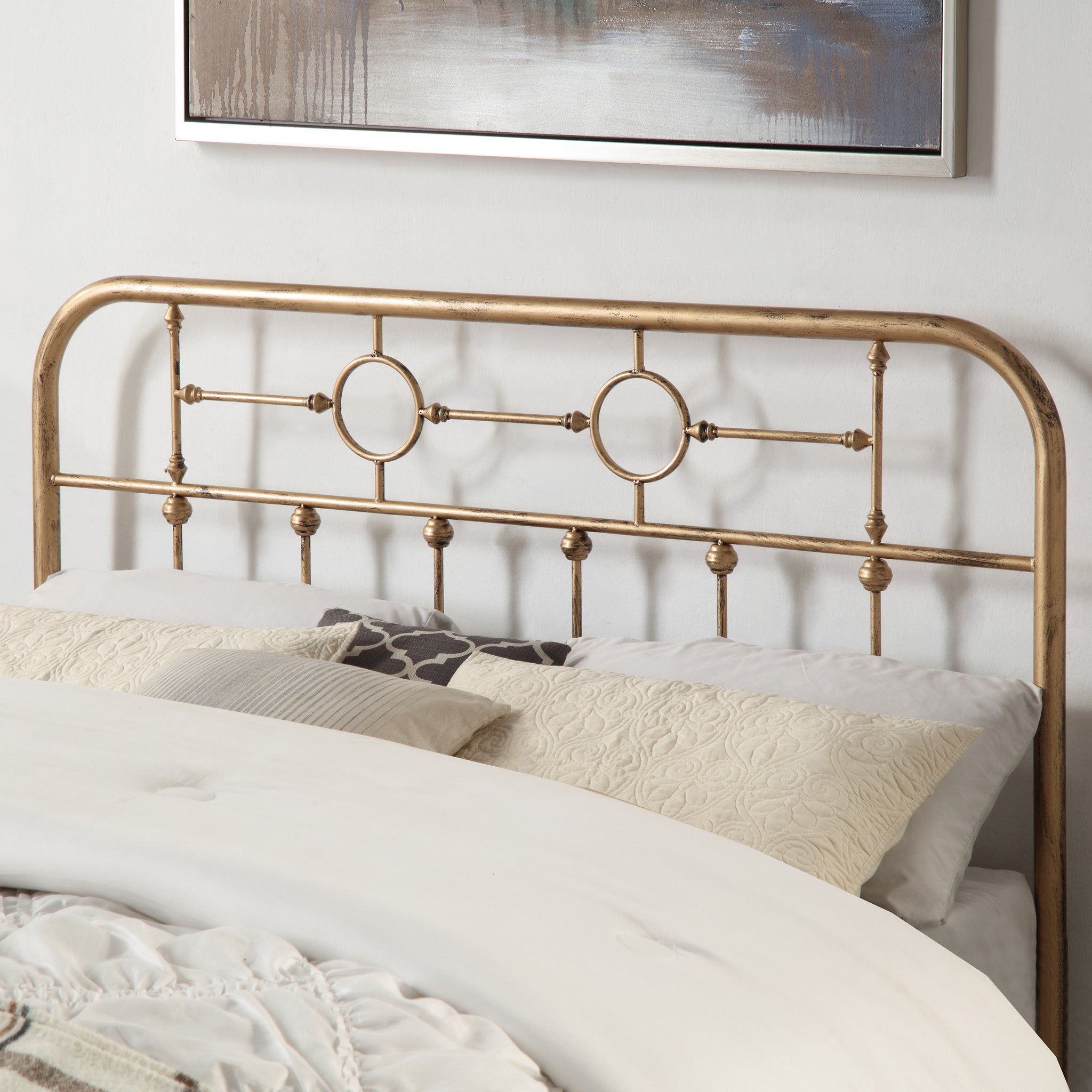 Brass-coloured metal headboard with circular designs and vertical bars, shown with decorative pillows. A painting hangs on the headboard.