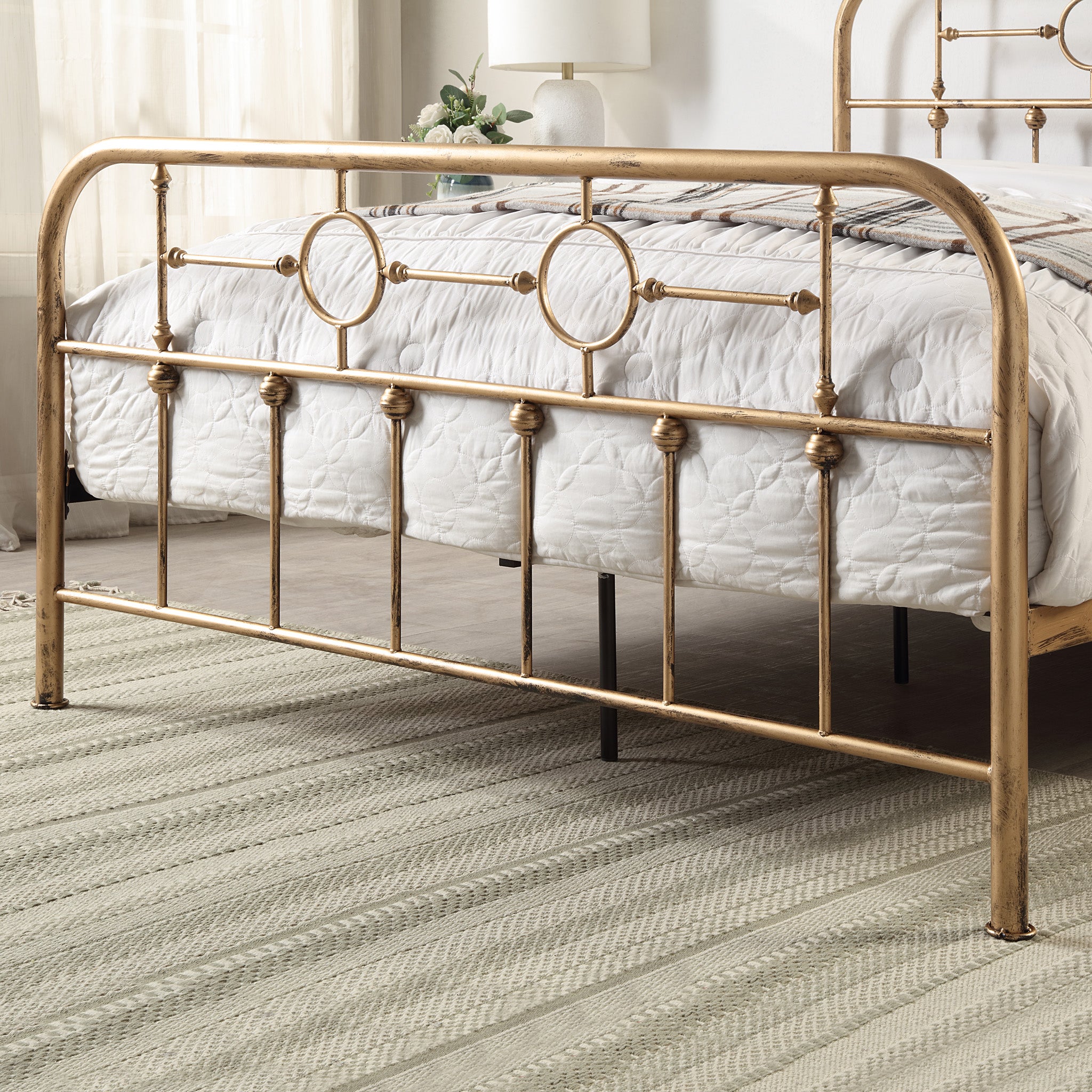 Brass-coloured metal bed frame with circular designs and vertical bars at the foot of a bed with a white quilt, and a beige carpet and white lamp in the background.