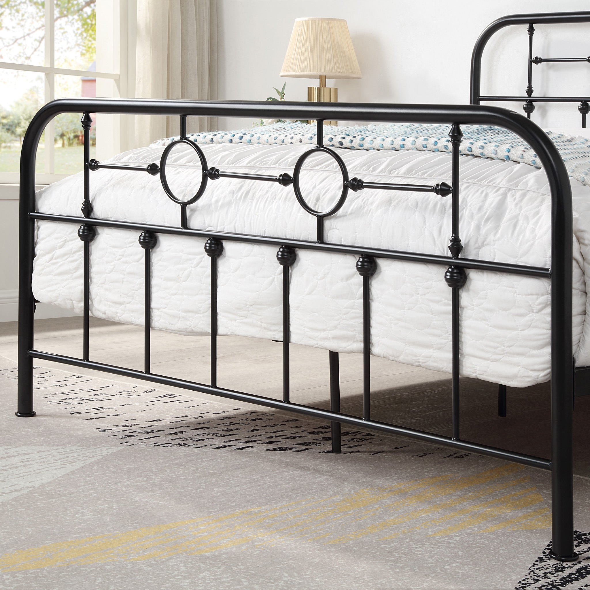 Close-up of a black metal bed frame with circular designs and vertical bars at the foot of the bed. White bedding with a patterned throw is visible. A lamp and window are in the background.