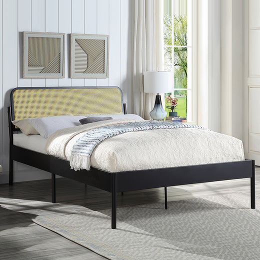 Black metal bed with a woven rattan headboard and quilted beige bedding. The room features white paneled walls, framed geometric artwork, a bedside table with a lamp, and a window with curtains.