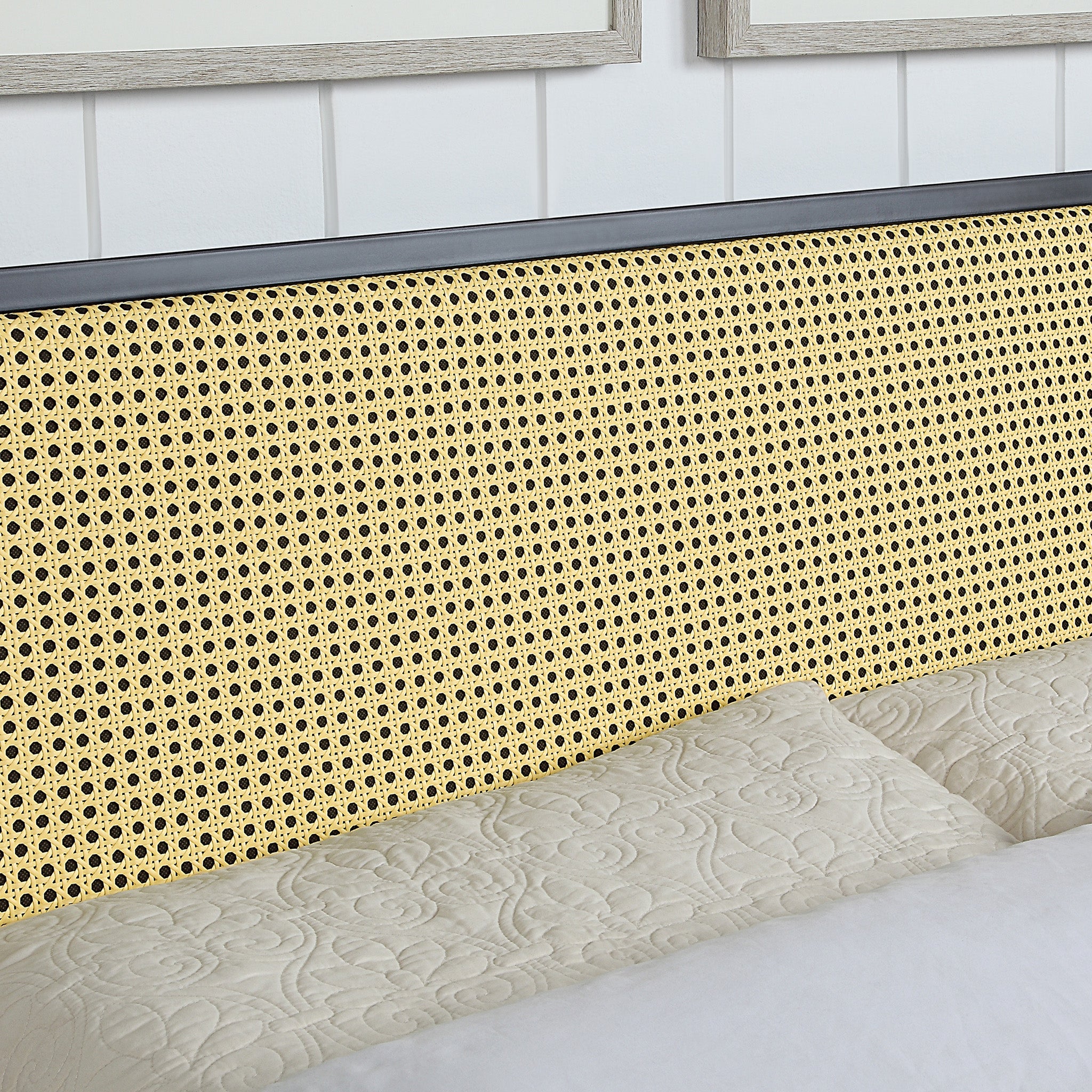 Close-up of a bed with a woven rattan headboard. The bed is made with quilted beige bedding, and the background features white paneled walls with framed artwork.