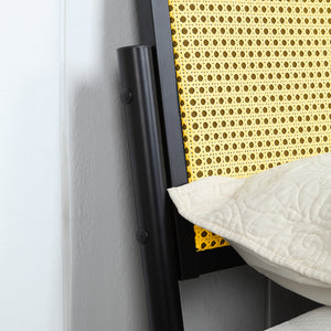 Close-up of a black metal bed frame with a woven rattan headboard. A beige quilted pillow is partially visible against the headboard. The background shows a plain white wall.