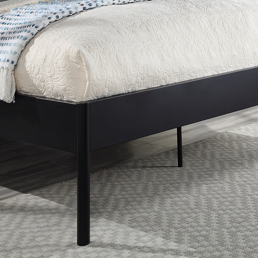Close-up of the corner of a black metal bed frame with quilted beige bedding and a blue and white throw blanket. The bed is on a light gray carpet, and the floor is dark wood.