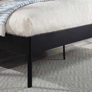Close-up of the corner of a black metal bed frame with quilted beige bedding and a blue and white throw blanket. The bed is on a light gray carpet, and the floor is dark wood.