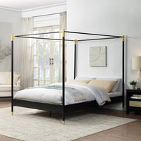 A black metal four-poster bed with gold accents on the corners and feet, set against a white wall, beside a large window with light curtains.