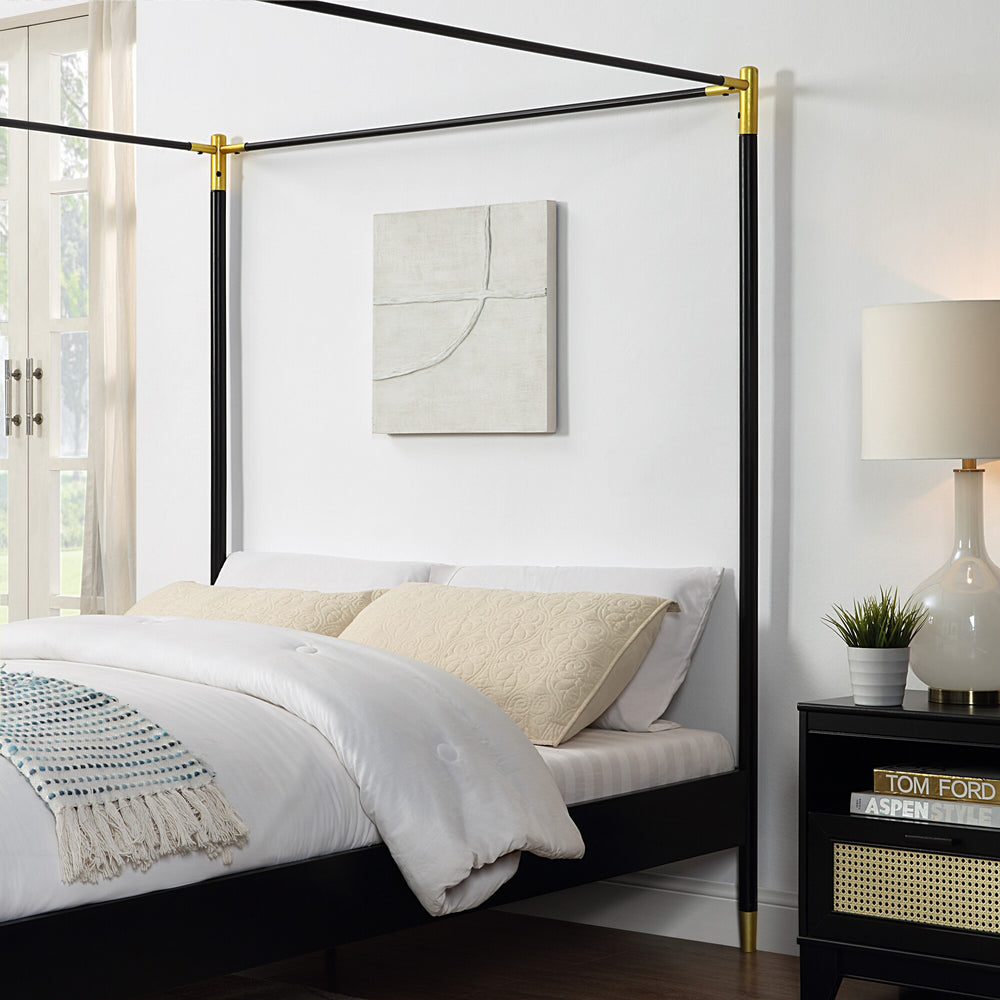A close-up of a black metal four-poster bed frame with gold accents, set against a white wall, next to a bedside table with a lamp and plant.