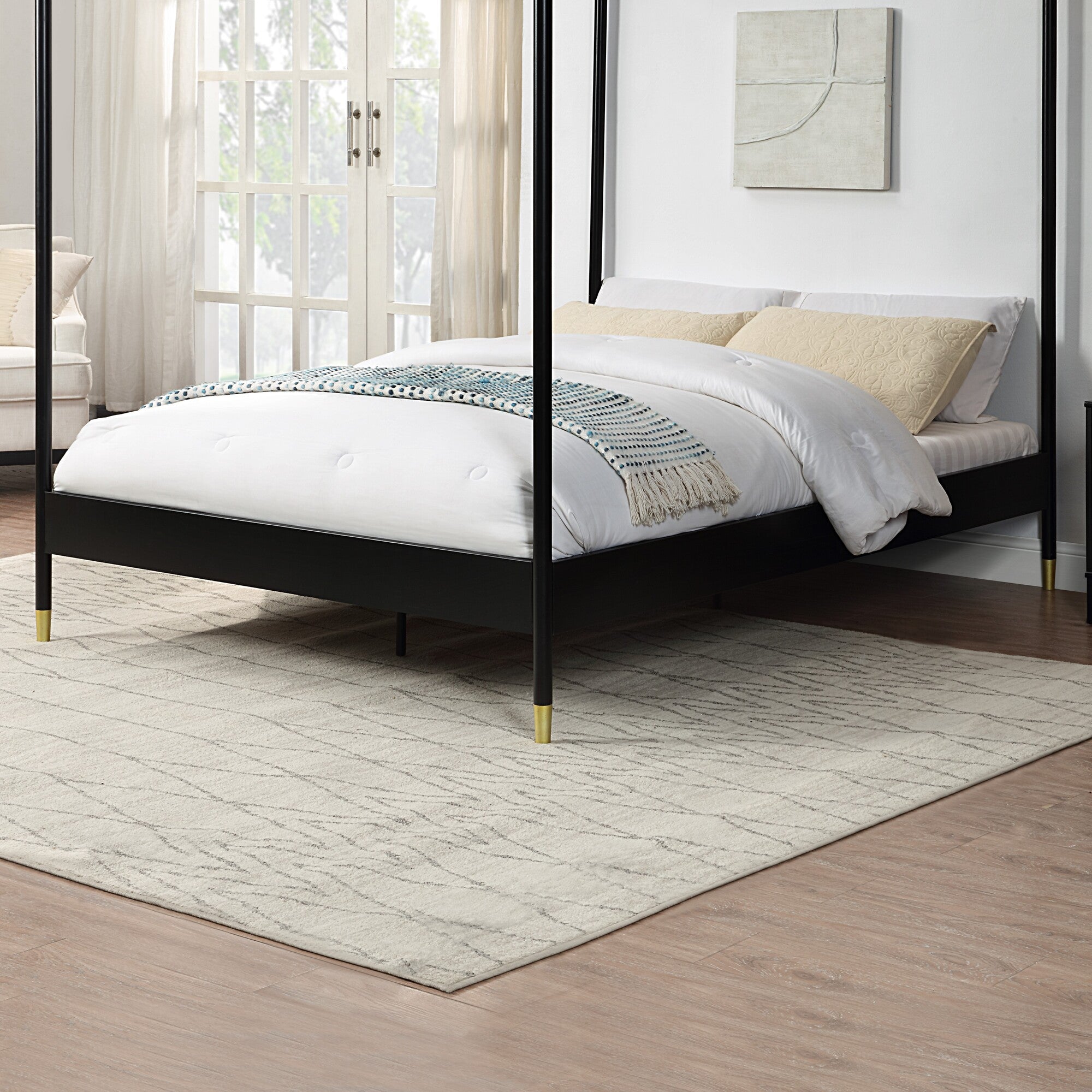 A black metal four-poster bed with gold accents on the feet, set on a light-coloured rug, beside a large window with light curtains.