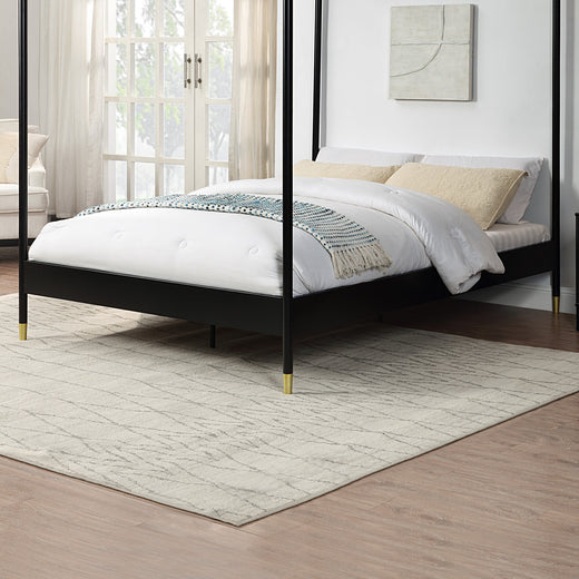 A black metal four-poster bed with gold accents on the feet, set on a light-coloured rug, beside a large window with light curtains.