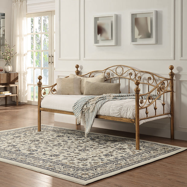 Bronze-coloured metal daybed with swirl design and finials, placed on a patterned rug in a bright room. Two abstract paintings hang on the white panelled wall above.