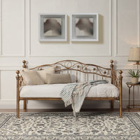 Bronze-coloured metal daybed with swirl design and finials, set against a white panelled wall on a patterned rug. Two abstract paintings hang above.