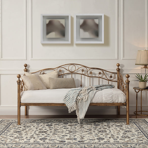 Bronze-coloured metal daybed with swirl design and finials, set against a white panelled wall on a patterned rug. Two abstract paintings hang above.