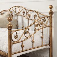 Close-up of the bronze-coloured metal daybed's side, featuring intricate swirl designs, floral accents, and finials, set against a white panelled wall.