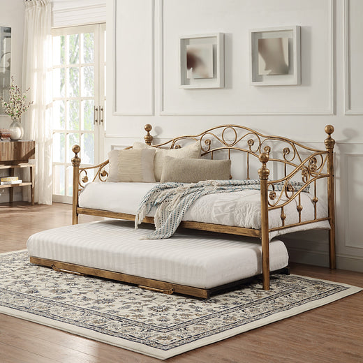 Bronze-coloured metal daybed with swirl design and finials, featuring a partially extended pull-out trundle, set against a white panelled wall on a patterned rug. Two abstract paintings hang on the wall above.