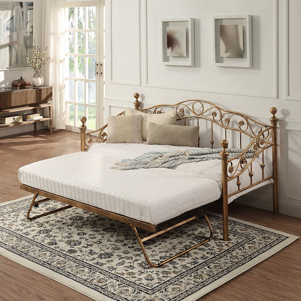 Bronze-coloured metal daybed with swirl design and finials, featuring an extended pull-out trundle, set against a white panelled wall on a patterned rug. Two abstract paintings hang on the wall above.