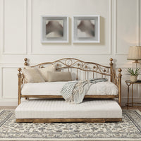 Bronze-coloured metal daybed with swirl design and finials, featuring a pull-out trundle, set against a white panelled wall on a patterned rug. Two abstract paintings hang on the wall above.