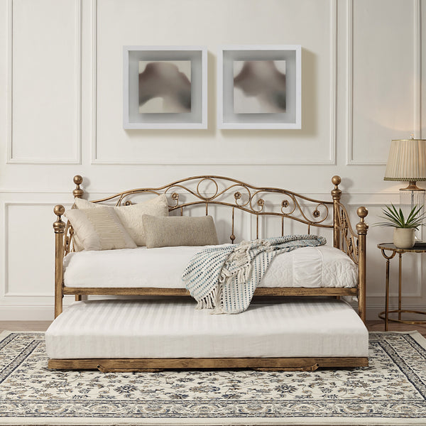 Bronze-coloured metal daybed with swirl design and finials, featuring a pull-out trundle, set against a white panelled wall on a patterned rug. Two abstract paintings hang on the wall above.