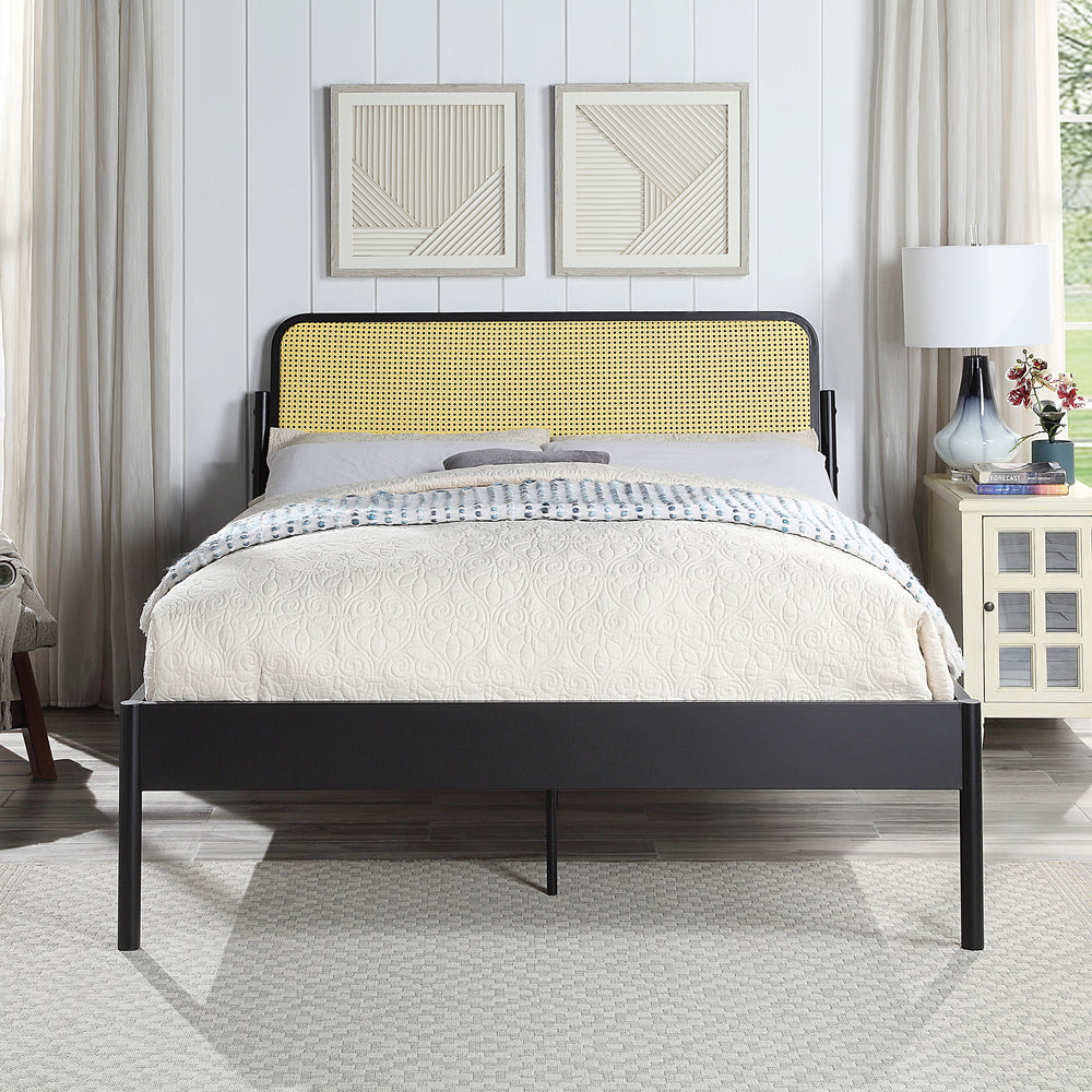 Black metal bed with a woven rattan headboard and quilted beige bedding. The room features white paneled walls, framed geometric artwork, a bedside table with a lamp, and a window with curtains.