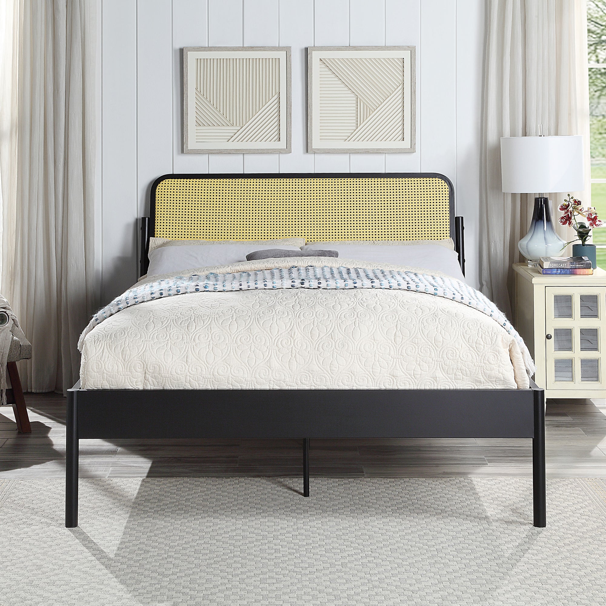 Black metal bed with a woven rattan headboard and quilted beige bedding. The room features white paneled walls, framed geometric artwork, a bedside table with a lamp, and a window with curtains.