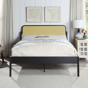 Black metal bed with a woven rattan headboard and quilted beige bedding. The room features white paneled walls, framed geometric artwork, a bedside table with a lamp, and a window with curtains.