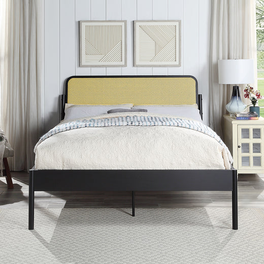 Black metal bed with a woven rattan headboard and quilted beige bedding. The room features white paneled walls, framed geometric artwork, a bedside table with a lamp, and a window with curtains.