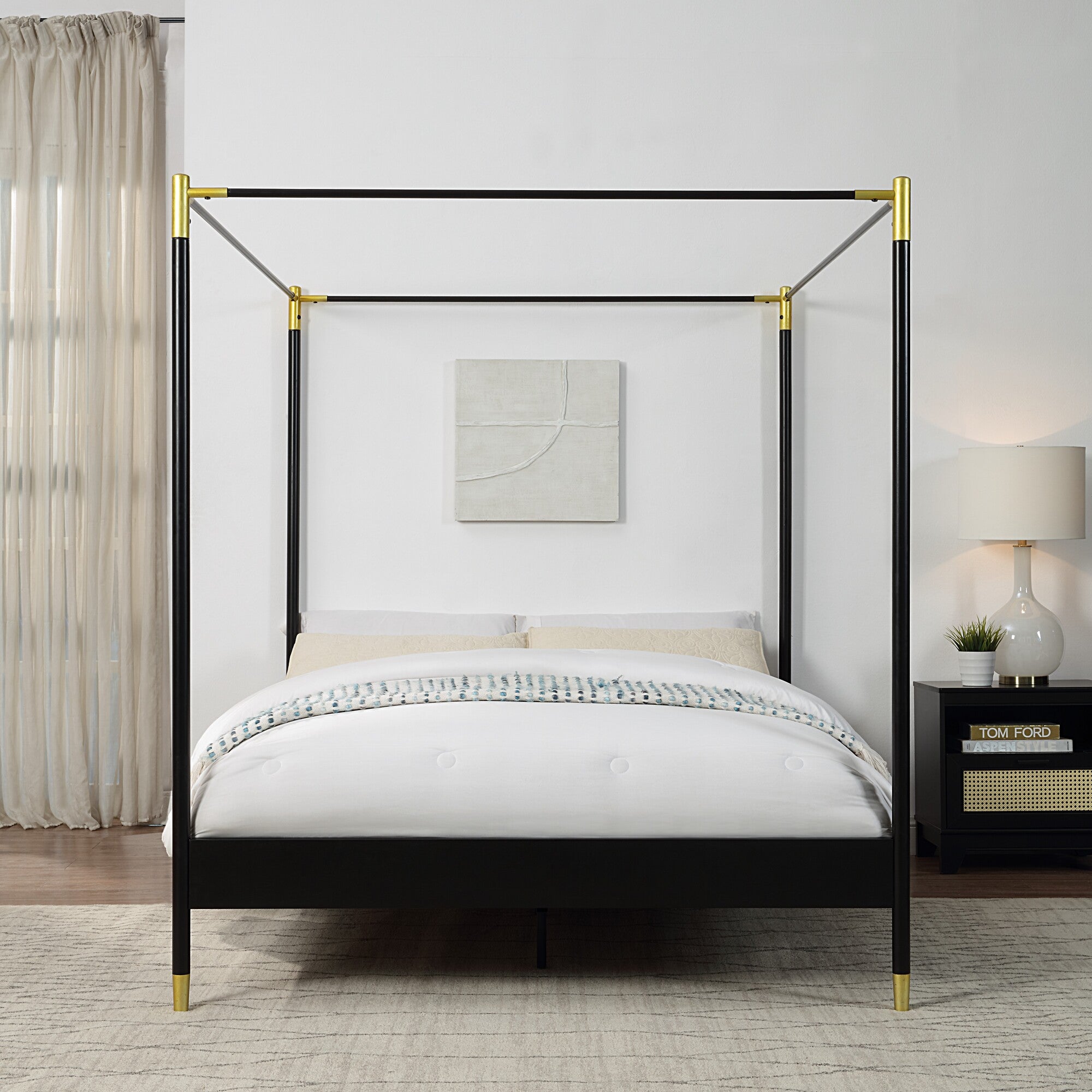 A black metal four-poster bed with gold accents on the corners and feet, set against a white wall, beside a large window with light curtains.