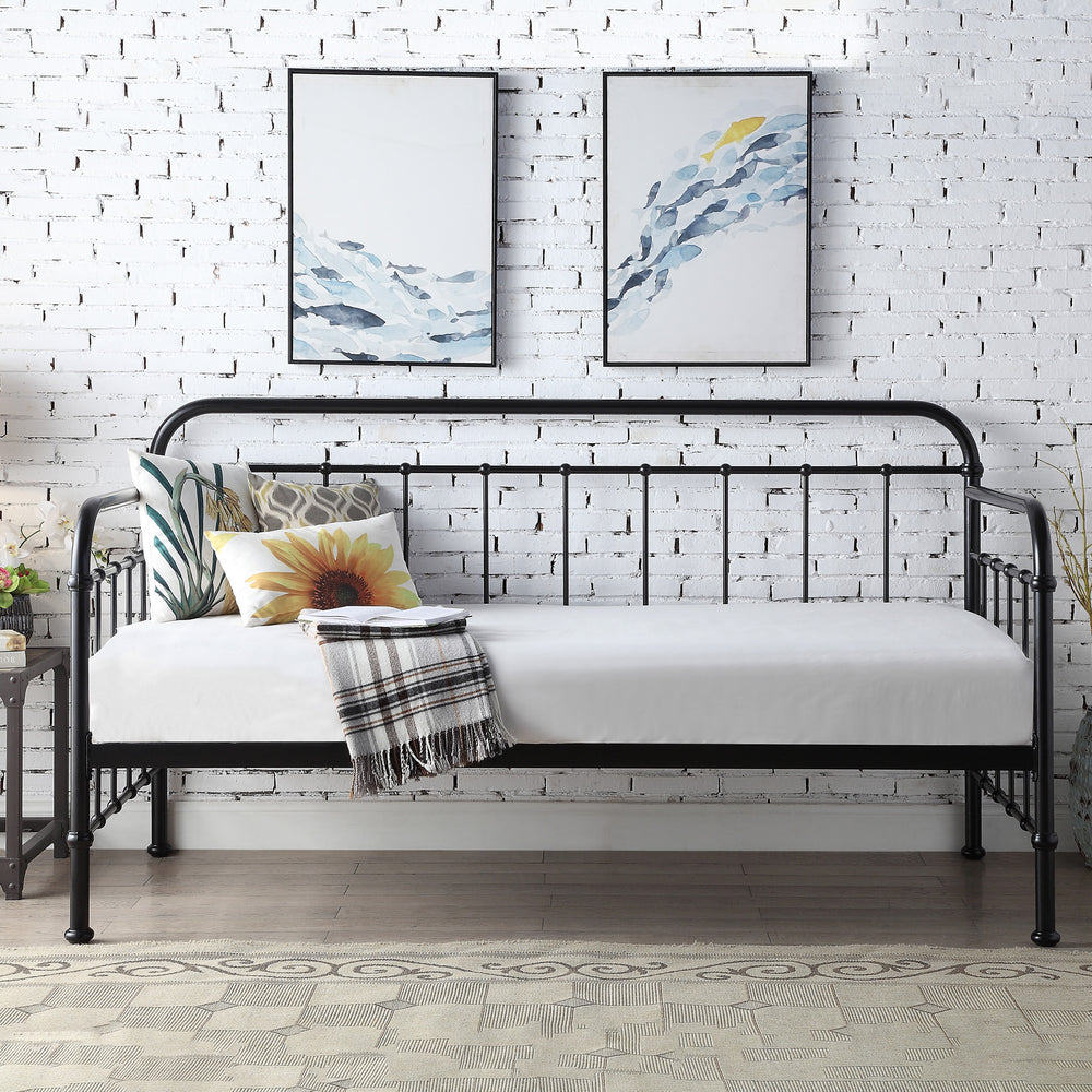 A black metal day bed is adorned with white bedding, decorative pillows, and a plaid throw blanket. Two paintings hang above the bed on a white brick wall.