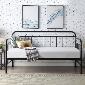 A black metal day bed is adorned with white bedding, decorative pillows, and a plaid throw blanket. Two paintings hang above the bed on a white brick wall.