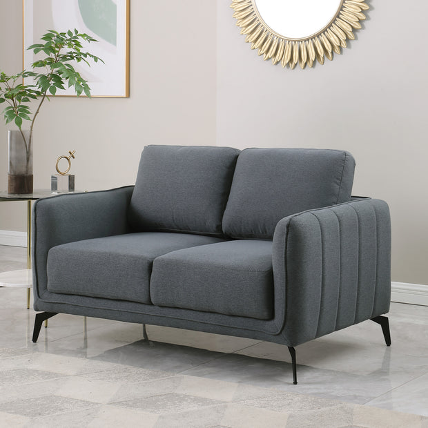 Dark grey fabric sofa with black legs, featuring vertical stitching on the armrests. The backdrop includes a stylish room with a decorative mirror, plant, and modern art.