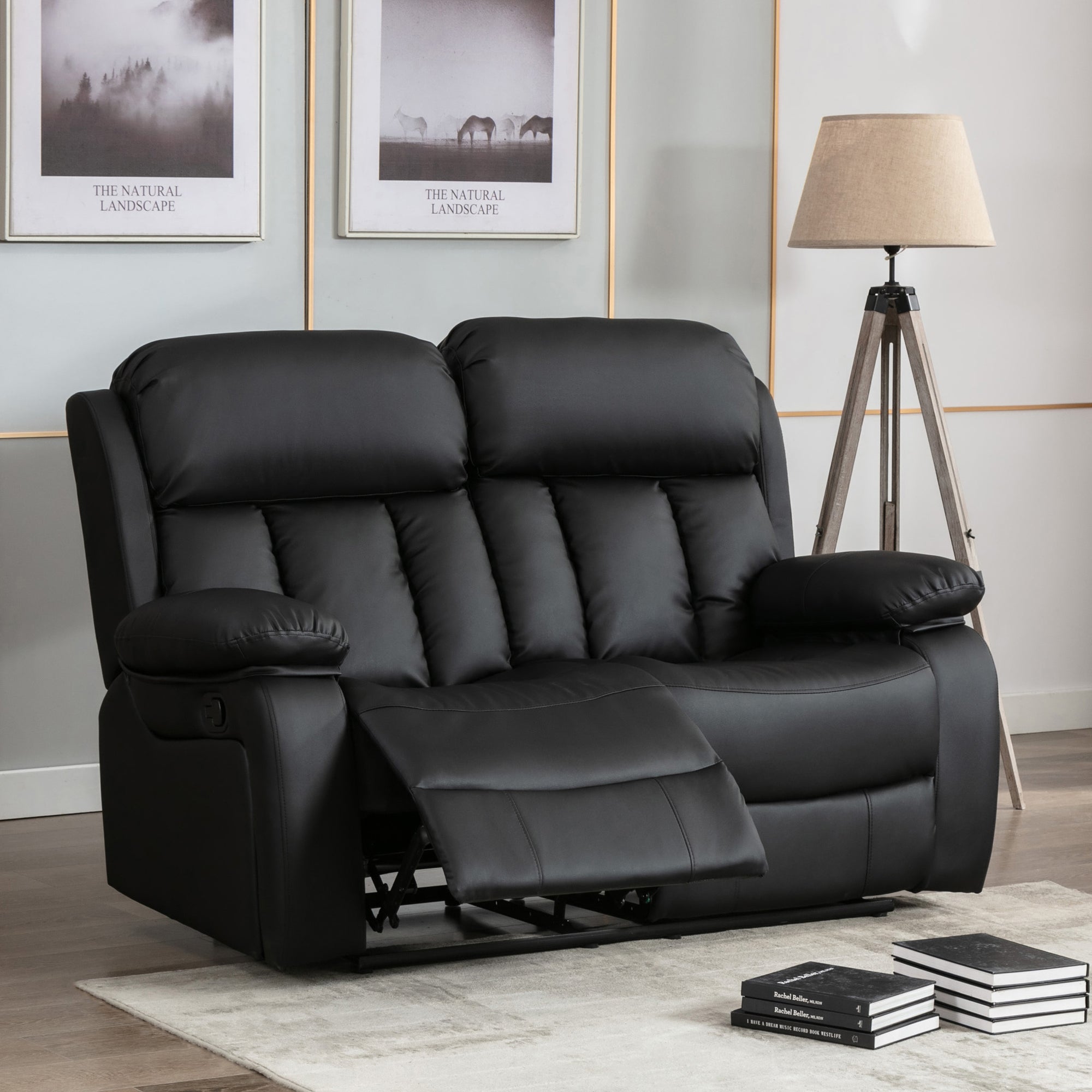 Black leather two-seater manual recliner sofa with padded armrests and vertically stitched backrests, with one seat reclined. The sofa is in a living room with a tripod floor lamp and landscape-themed wall art.