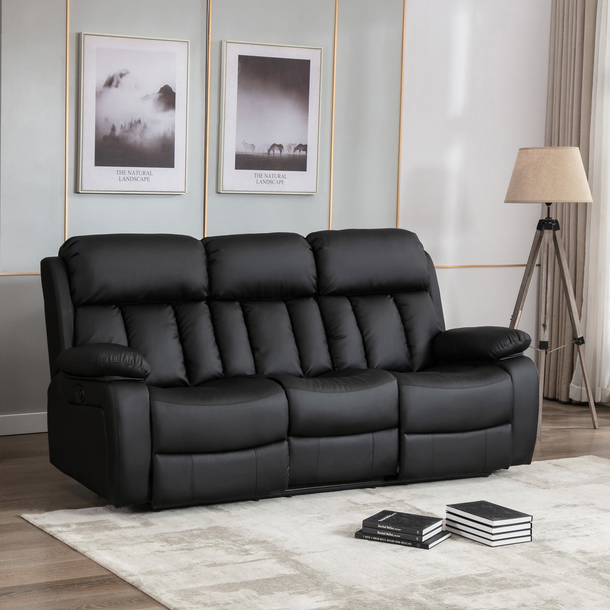 Black leather three-seater electric recliner sofa with padded armrests and cushioned backrests featuring vertical stitching, placed on a light-coloured rug in a living room. Two photos hang above the sofa.