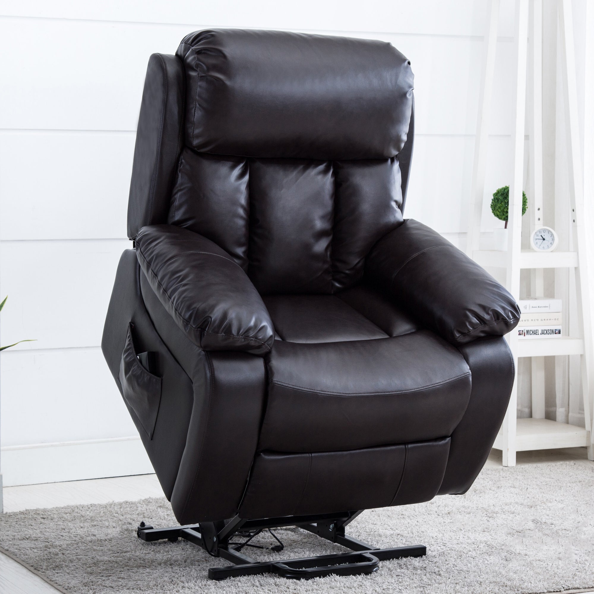 Black leather rise recliner chair on a grey rug in a bright living space, featuring a small white shelf with books and a plant in the background.