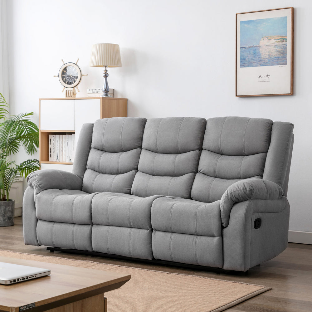Grey fabric three-seater electric reclining sofa in a cosy living room with light wooden furniture, a nautical clock, a table lamp, and framed artwork.