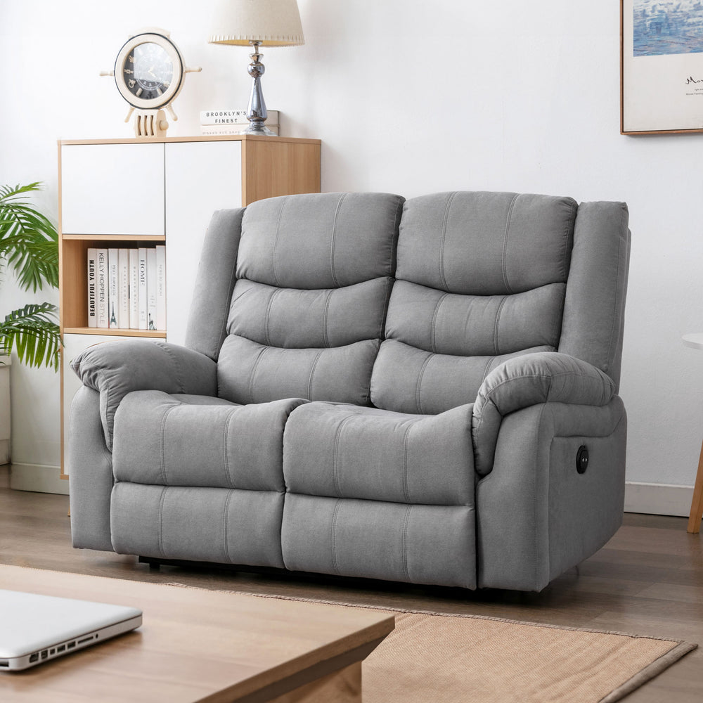 Grey fabric two-seater electric reclining sofa in a cosy living room with light wooden furniture, a nautical clock, a table lamp, and framed artwork.