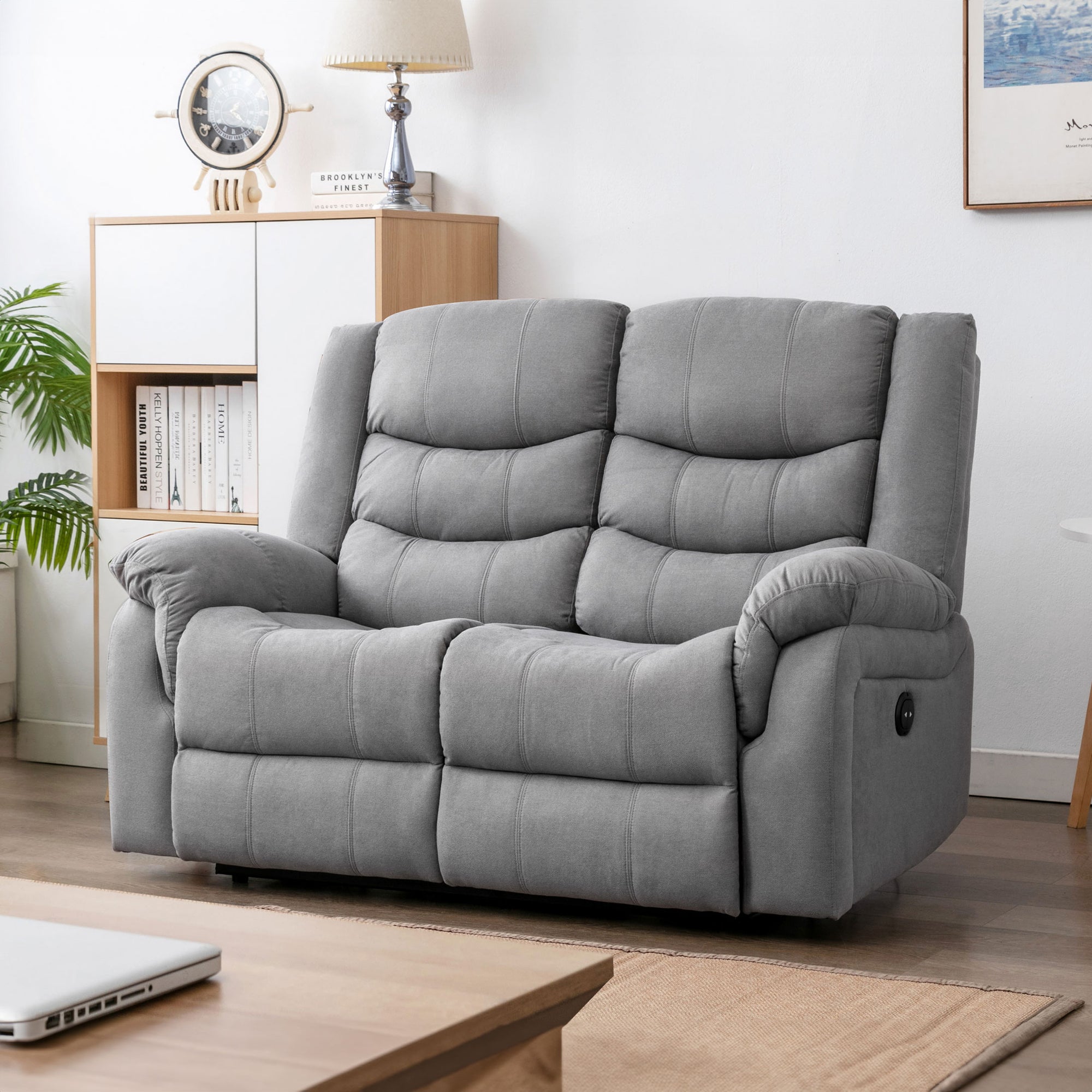 Grey fabric two-seater electric reclining sofa in a cosy living room with light wooden furniture, a nautical clock, a table lamp, and framed artwork.
