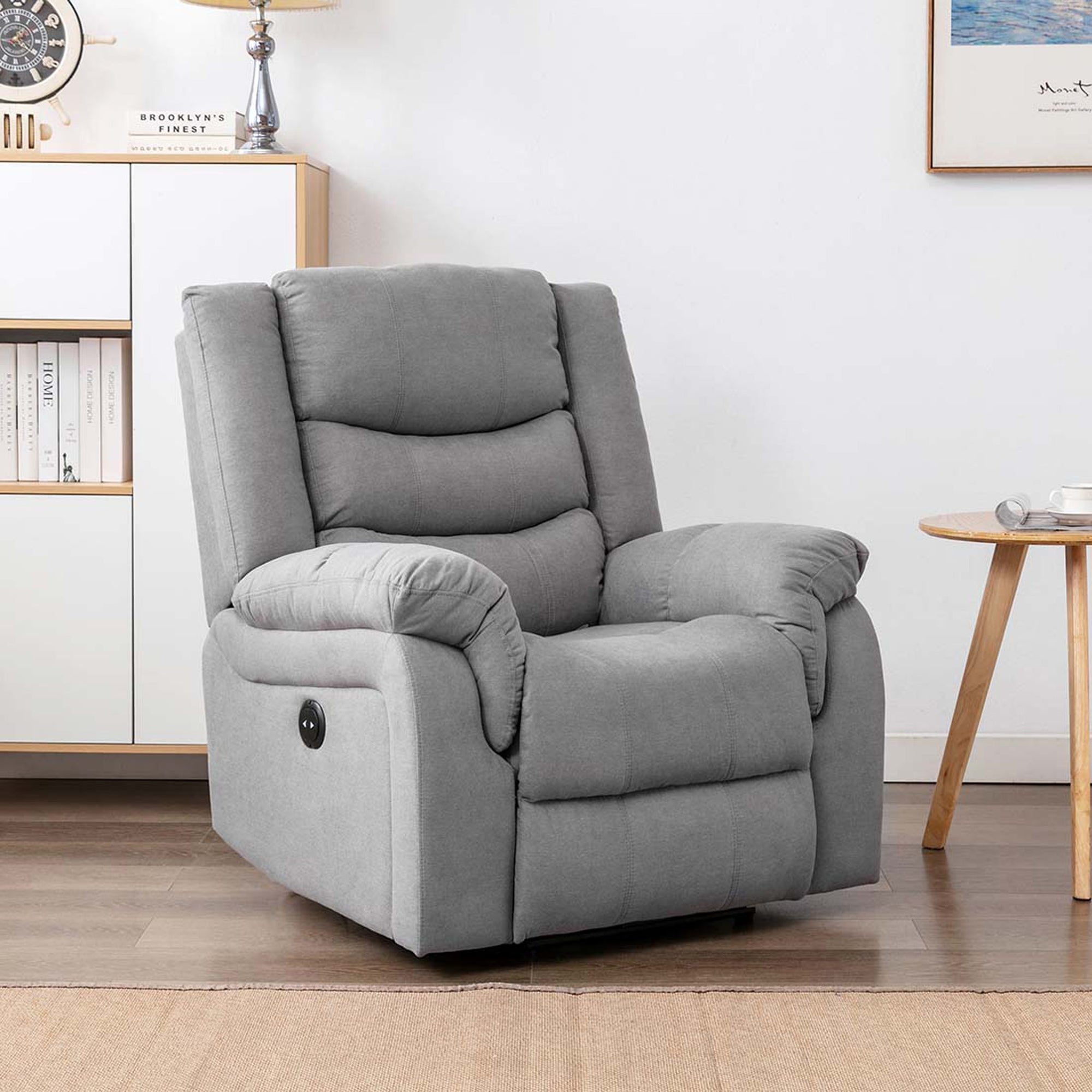 Grey fabric electric recliner chair in a cosy living room with light wooden furniture, a nautical clock, a table lamp, and framed artwork.