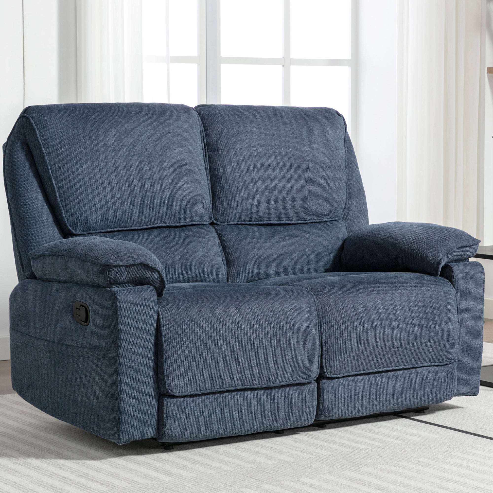 Blue fabric manual reclining two-seater sofa with padded armrests, placed on a light-coloured rug in a living room near large windows.