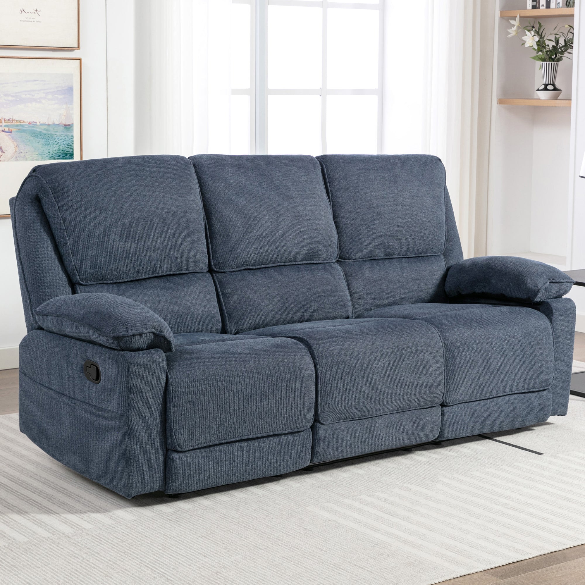 Blue fabric manual reclining three-seater sofa with padded armrests, positioned on a light-coloured rug in a living room near large windows.