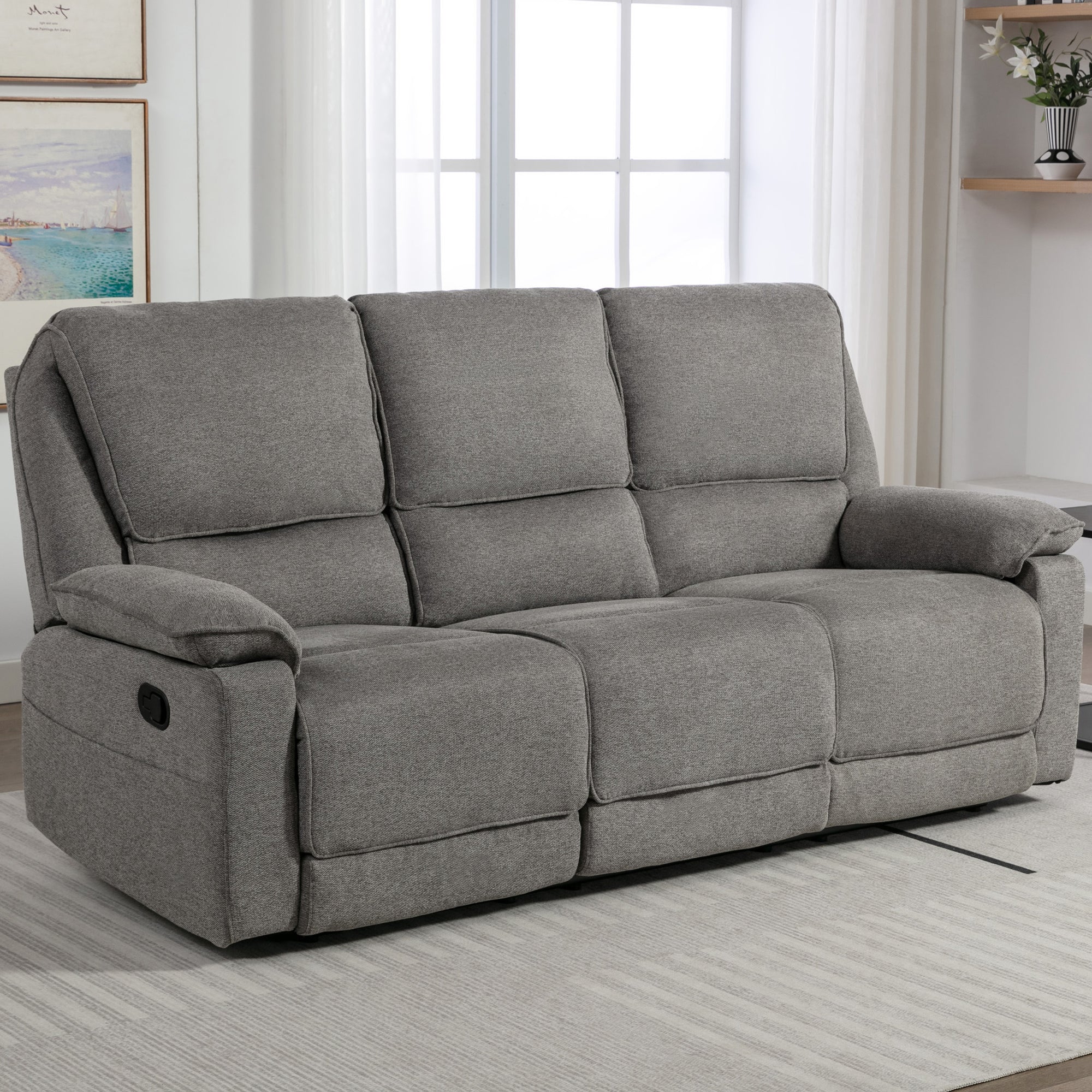 Grey fabric manual reclining three-seater sofa with padded armrests, positioned on a light-coloured rug in a living room near large windows.