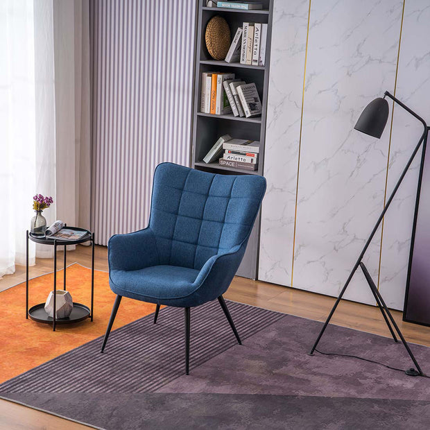 A blue linen accent chair with black tapered legs, placed in a room with a round side table, a floor lamp, and a bookshelf filled with books and decorative items.