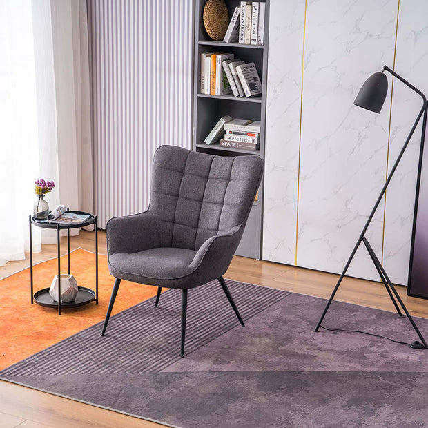 A charcoal linen accent chair with black tapered legs, placed in a room with a round side table, a floor lamp, and a bookshelf filled with books and decorative items.