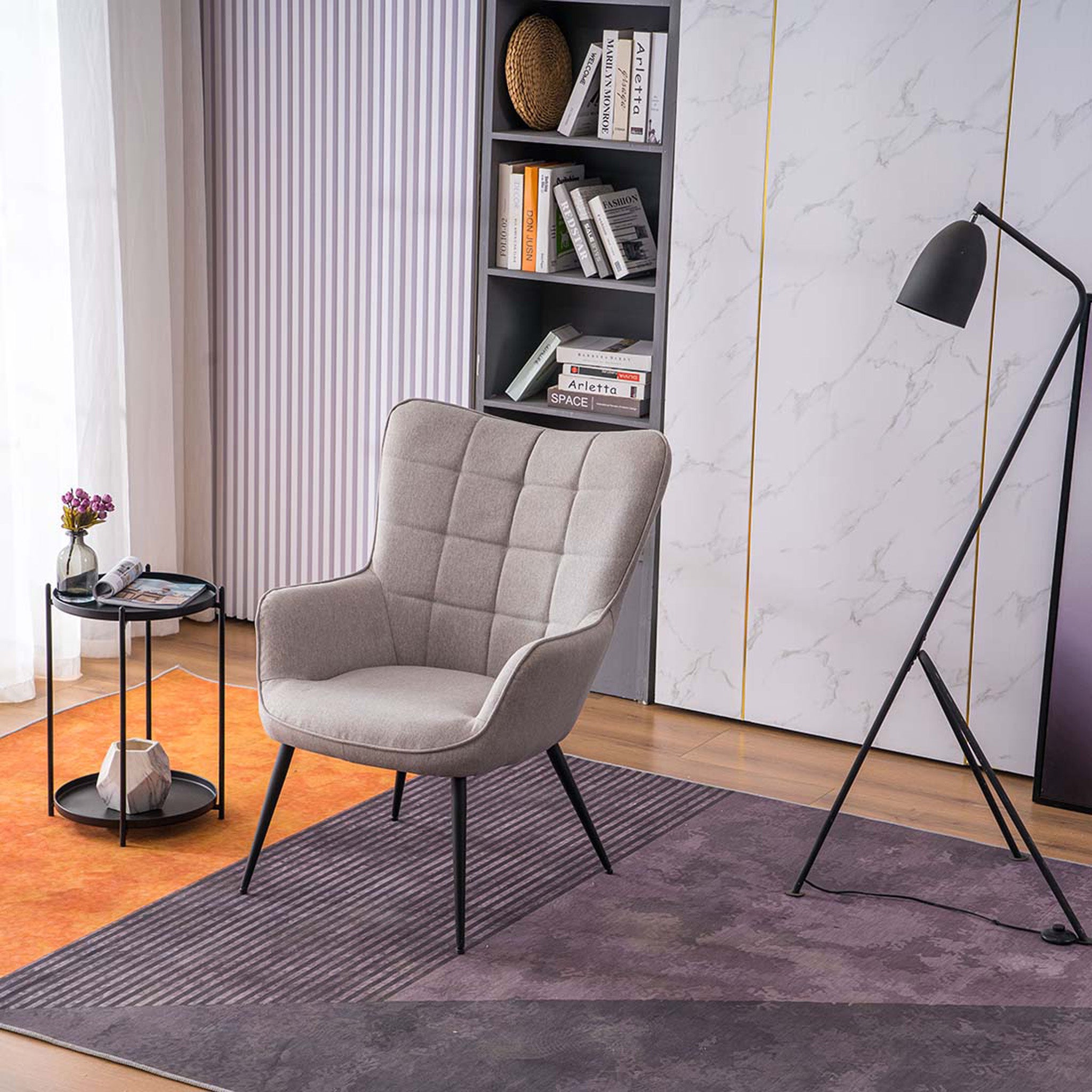 A light grey linen accent chair with black tapered legs, placed in a room with a round side table, a floor lamp, and a bookshelf filled with books and decorative items.