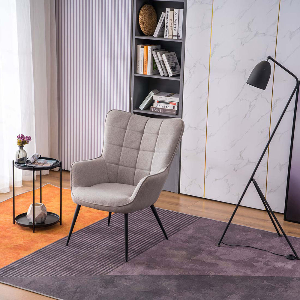 A light grey linen accent chair with black tapered legs, placed in a room with a round side table, a floor lamp, and a bookshelf filled with books and decorative items.