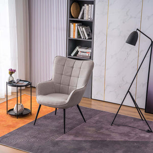 A light grey linen accent chair with black tapered legs, placed in a room with a round side table, a floor lamp, and a bookshelf filled with books and decorative items.