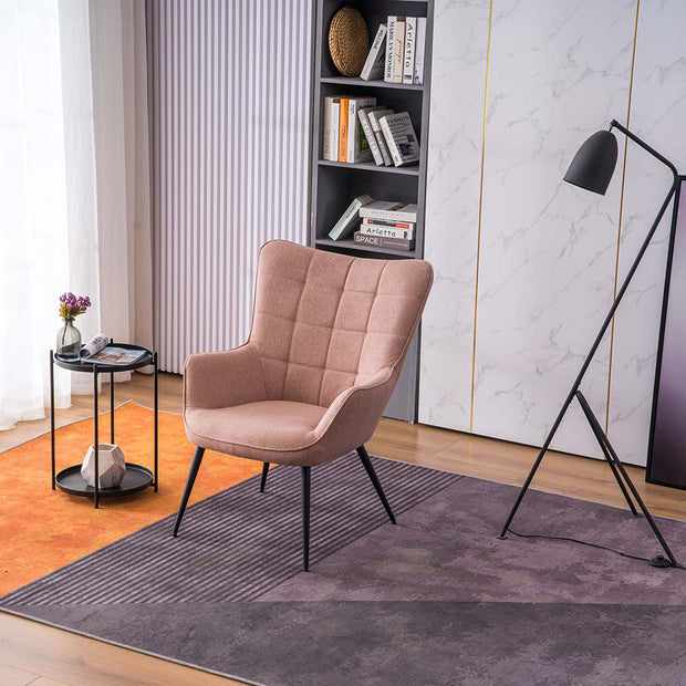 A pink linen accent chair with black tapered legs, placed in a room with a round side table, a floor lamp, and a bookshelf filled with books and decorative items.