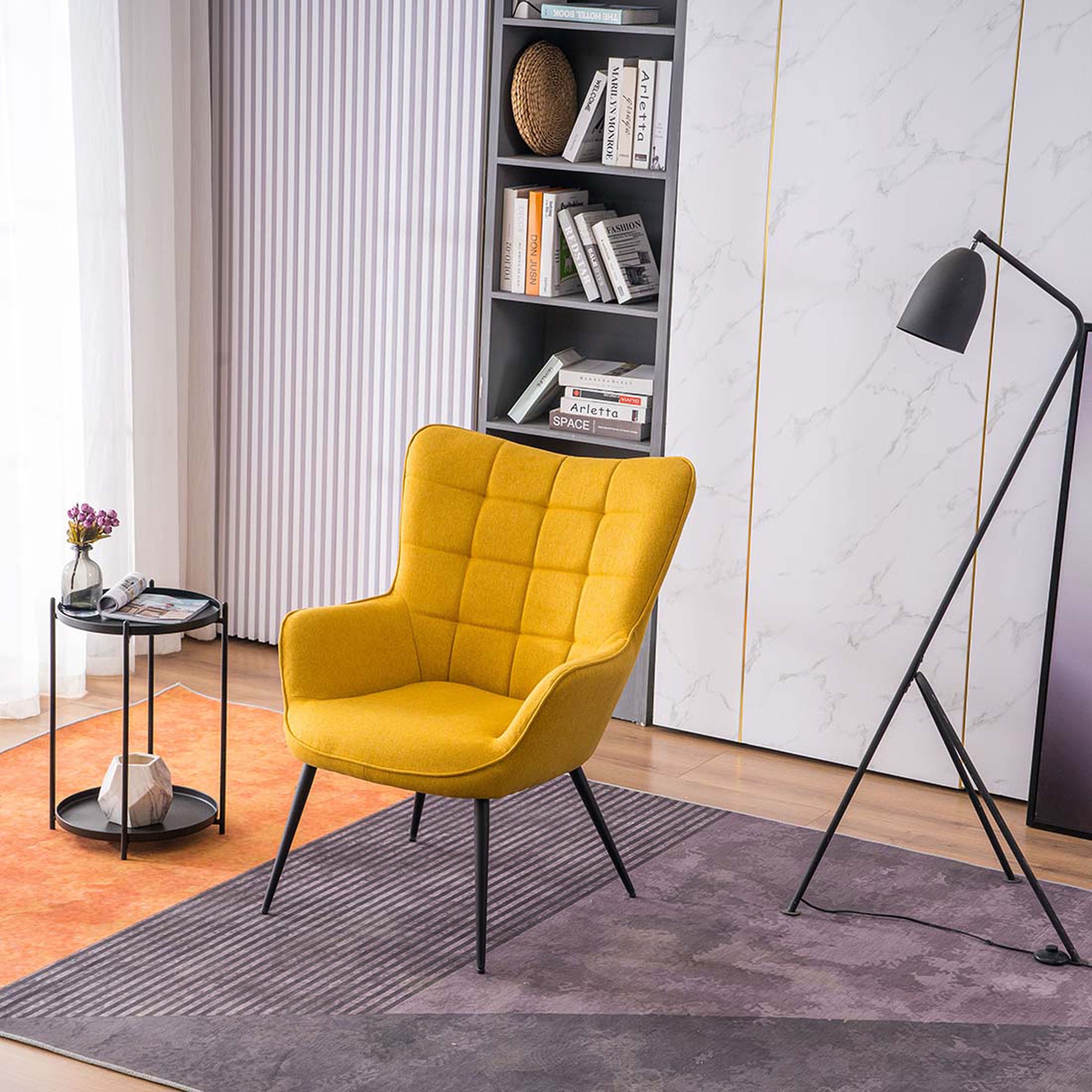 A yellow linen accent chair with black tapered legs, placed in a room with a round side table, a floor lamp, and a bookshelf filled with books and decorative items.